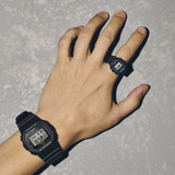 A hand wears a black digital wristwatch, the CASIO G-Shock Watch nano SERIE 5600 by G-Shock Watches, along with a matching digital ring watch styled like a G-Shock Nano, both displayed against a textured gray background.