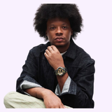 A person with an afro hairstyle sits cross-legged, dressed in a dark denim jacket and light pants, wearing the G-Shock Watches CASIO G-Shock Watch Bold Camo Series GA700CMG-3A. They look directly at the camera with a neutral expression against a plain light background.