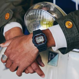 A person in a green jacket with large brown buttons rests their hands near a round headlight of a vintage vehicle, wearing the G-Shock Watches CASIO G-Shock Watch DIGITAL DW5000R-1A in black.