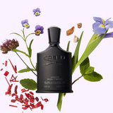 A black bottle of Creed Green Irish Tweed 3.4 EDP Men Perfume by Creed Perfumes sits among purple flowers, green leaves, wood shavings, and small stones on a light background.