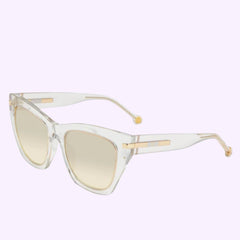 The Carolina Herrera SHE831V Women Sunglasses feature clear cat-eye frames, light brown gradient lenses, and gold accents on the temples, showcased against a light purple background.