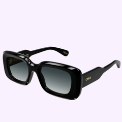 Discover the Chloe CH0188S 001 Woman Recycled Acetate Sunglass by Chloe Sunglasses—a pair of black rectangular sunglasses with dark gradient lenses and distinctive gold Chloé branding on the bold arms. Expertly crafted from recycled acetate, these designer women’s sunglasses offer a blend of sustainability and luxury. Displayed against a light purple background, they exemplify chic, eco-conscious style.