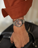 A person wearing a Citizen Watches CA4664-60X Promaster Eco-Drive, a chronograph stainless steel wristwatch with a pink and black dial by Citizen Watches (UPC: 013205169588), is holding the handle of a black leather bag. The distinctive timepiece stands out against their rust-colored long-sleeve shirt.