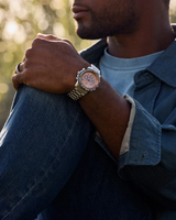 A man sits outdoors with his arm resting on his knee, wearing a blue denim shirt and a Citizen Watches CA4664-60X Promaster Eco-Drive chronograph watch (UPC: 013205169588) by Citizen Watches, which features a brown face. The background is blurred greenery.