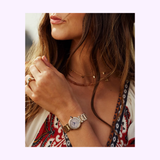 A woman with long brown hair wears a Citizen Watches EM1136-87D L Mae gold women's watch, layered necklaces, and a red-and-white patterned top. She holds her hand near her chin, displaying a ring on her finger.