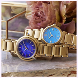 Certainly! Here’s a revised version of the sentence, incorporating the provided product data: Two gold Citizen Watches rest on a marble surface—one is the elegant Citizen Watches L Mae EM1133-85L Limited Edition, an Eco-Drive ladies’ watch with a deep blue face; the other features a light blue face. Both limited edition watches are framed by blurred flowers and a glass box in the background.