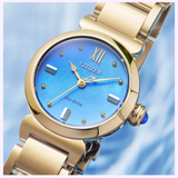 The Citizen Watches L Mae EM1133-85L Limited Edition is a gold ladies’ watch by Citizen Watches, featuring a striking blue face with Roman numeral XII and luminous hour markers, showcased against a soft blue background.