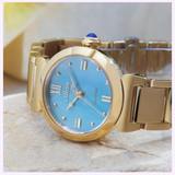 A Citizen Watches L Mae EM1133-85L Limited Edition ladies’ watch in gold with a blue dial rests on a light stone surface. This exclusive timepiece by Citizen Watches features silver hour markers, a gold band, and is accented by a petite blue jewel set into the crown. The softly blurred background draws attention to the elegant details of the watch.