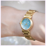A gold wristwatch with a blue face and gold Roman numeral markers is worn on a person’s left wrist. Reminiscent of the elegance found in limited edition timepieces, the watch brand Citizen Watches is visible on the dial. This timepiece is the Citizen Watches L Mae EM1133-85L Limited Edition.
