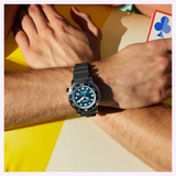 A wrist wearing a Citizen Watches Promaster Dive BN0167-09W with a patterned blue dial and black silicone strap, with a playing card visible in the background.