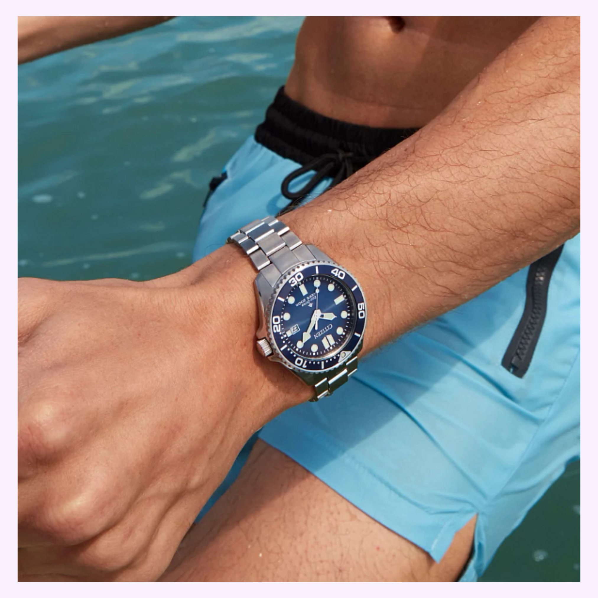 A person in blue swim trunks stands by the water, showcasing their Citizen Watches Promaster Dive BN0260-54L with a stainless steel band and blue face in the foreground.