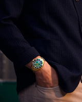 A person wearing a dark textured jacket and light pants has their hand in their pocket, revealing a Citizen Watches Promaster Dive BN0262-59W on their wrist—a gold-tone stainless steel timepiece with a green face and bezel from Citizen Watches.