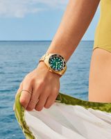 A person in a yellow swimsuit stands by the ocean, wearing the Citizen Watches Promaster Dive BN0262-59W by Citizen Watches—a gold-tone stainless steel Eco-Drive watch with a green face—and holding a white-and-green cloth.