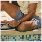 A person in light blue shorts and a cream knit shirt, wearing a Citizen Watches Promaster Dive LIGHT in BLACK BN0264-53E, sits cross-legged by a tiled pool, holding open a magazine.