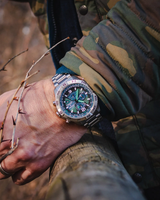 A person wearing a camouflage jacket and a Citizen Watches Promaster Navihawk A-T AT8220-55W, featuring a green and black dial, rests their hand on a wooden fence outdoors.