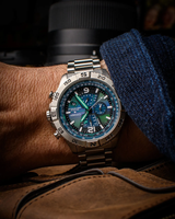 A close-up of a Citizen Watches Promaster Navihawk A-T AT8220-55W, a silver chronograph wristwatch with a striking blue and green dial featuring advanced Atomic Timekeeping, worn on a person’s wrist, with part of a camera lens and a brown leather bag in the background.