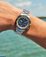 A close-up of a person’s wrist wearing the Citizen Watches Promaster Navihawk CA4667-53L, a silver chronograph watch by Citizen Watches featuring a blue and yellow bezel, photographed outdoors with water and sky softly blurred in the background.
