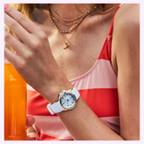 A woman in a striped bikini holds a drink, wearing the Citizen Watches Veya Silver FE1262-03A and layered necklaces with a crescent moon pendant.