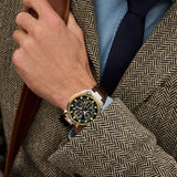 Citizen Watches Promaster Tsuno Chrono Racer AV0104-06W with a green face and gold accents, worn on the wrist of a person in a brown herringbone blazer and blue dress shirt.