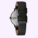 The Caravelle Watches By Bulova 44A119 Min Max dress watch features a round silver case, black crown, and black leather strap with an engraved buckle. Displayed from the back against a light lavender background.