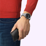 A person in a red long-sleeve shirt and blue jeans holds a black case, displaying the Tissot Watches PR516 Powermatic 80 (T1494071105100, UPC:7611608318492) automatic watch on their wrist.