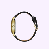 Side view of the Bulova 44B118 Dress Caravelle watch by Caravelle Watches By Bulova, showing its gold case, black leather strap, and clasp against a light purple background.