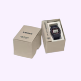 A CASIO G-Shock Watch DIGITAL DW5000R-1A by G-Shock Watches is partially displayed inside an open beige box, with a closed G-Shock branded box beside it on a light background.