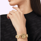 A person in a black textured sweater rests their hand on their chin, highlighting the Versace Watches VE3C00222 Stud Icon Bracelet Watch with chunky gold round links and an engraved design against a plain light background.