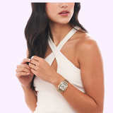A woman in a sleeveless white dress with a crisscross neckline showcases her Michele MWW06A000780 Deco 18k Gold Diamond Dial Watch by Michele Watches, featuring a gold rectangular face, as she adjusts her fingers. Her long, dark hair is styled in soft waves.