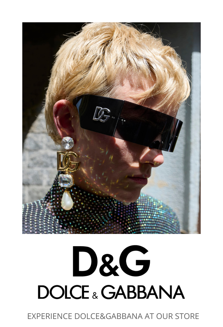 A person with short blonde hair wears oversized D&G sunglasses, a large jeweled Dolce & Gabbana earring, and a sparkling top. The D&G logo and text, “EXPERIENCE DOLCE&GABBANA AT OUR STORE,” appear below.