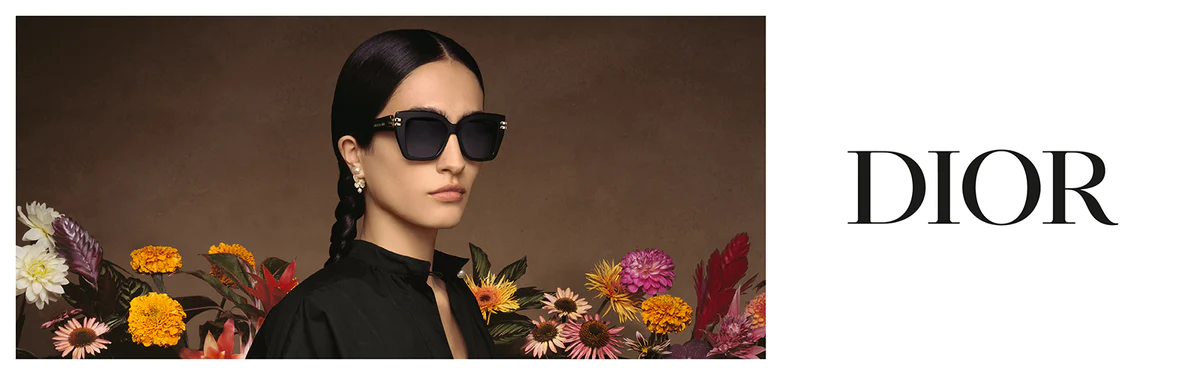 A woman with dark hair in braids wears large black sunglasses and statement earrings, dressed in black, standing in front of colorful flowers. The word DIOR appears in bold letters on the right.
