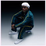 A man in a teal tracksuit with yellow stripes, white sneakers, and a white beret wears Dita Sunglasses LSA-404 DLS404-56-01RO featuring polarized lenses as he squats against a dark gradient background.