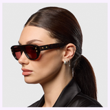A woman with straight, dark brown hair in a low ponytail wears Dita Sunglasses RHYTHX DTS741-A-03 Limited Edition navigator frames, silver hoop earrings, and a black jacket, shown in profile against a plain light background.
