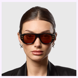 A woman with slicked-back dark hair wears Dita Sunglasses RHYTHX DTS741-A-03 Limited Edition, featuring a sculptural navigator shape and reddish lenses, paired with silver hoops and a black blazer, against a light grey background.