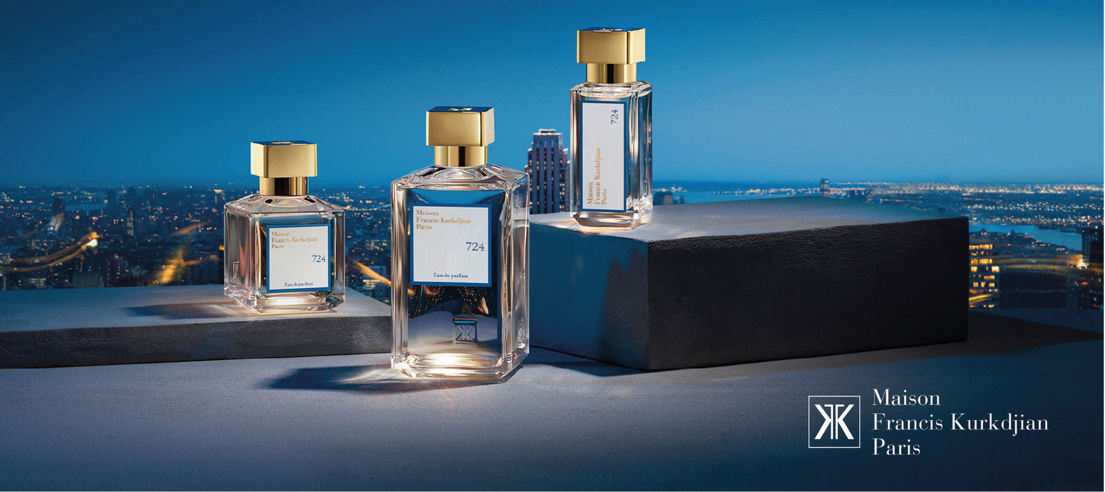 Three Maison Francis Kurkdjian Paris 724 perfume bottles, each with a gold cap, are displayed on platforms with a city skyline and evening sky in the background. The brand logo and name appear in the lower right corner.