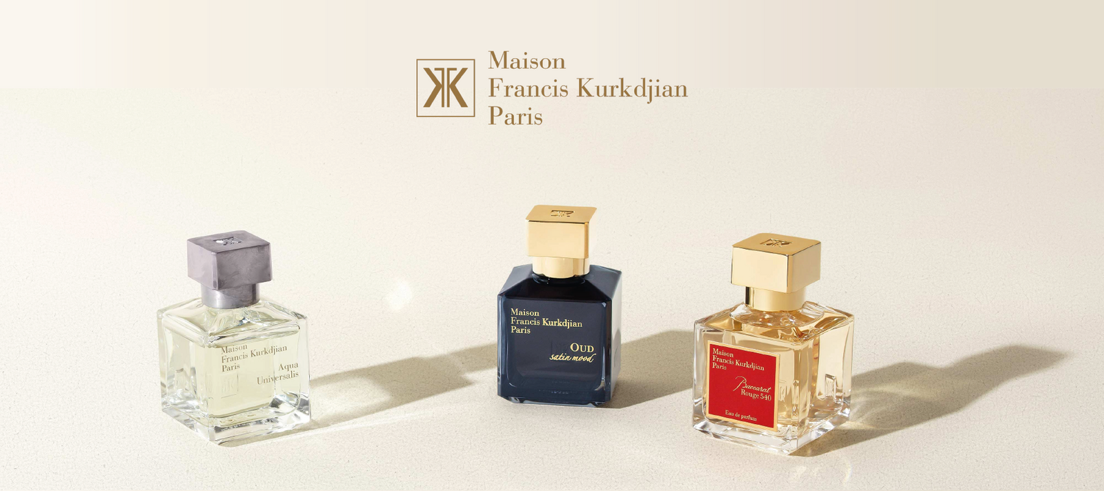 Three Maison Francis Kurkdjian Paris perfume bottles—clear, dark blue, and amber—are displayed on a light surface under a beige background with the brand’s logo and name at the top.
