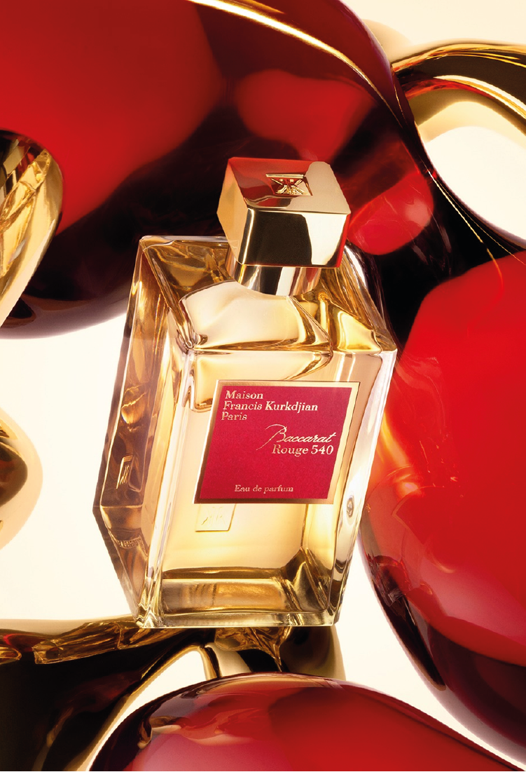 A rectangular glass bottle of Maison Francis Kurkdjian Baccarat Rouge 540 perfume is surrounded by shiny abstract shapes in red and gold. The perfume label is red with gold text, and the cap is metallic gold.