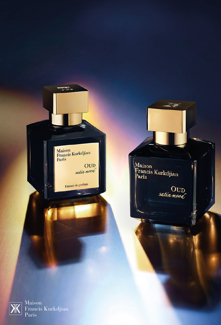Two dark glass perfume bottles with gold square caps, labeled Maison Francis Kurkdjian Paris OUD and OUD satin mood, are displayed on a reflective surface with soft, colorful lighting. Brand logo appears at the bottom left.