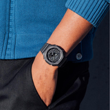 A person dressed in a blue sweater and black pants stands with their hand in their pocket, displaying the rugged G-Shock GA2100-1A1 2100 Series wristwatch (UPC: 889232241630) from G-Shock Watches—renowned for its impressive shock resistance—on their left wrist.