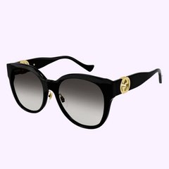 Gucci GG1028SK 006 unisex sunglasses from Gucci Sunglasses feature a sleek black cat-eye design, gradient gray lenses, and gold logo accents on the arms, showcased against a pale purple background.