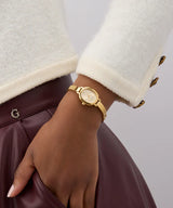 Wearing a Guess Watches GW0924L2 gold-tone watch, a white textured jacket with brown buttons, and resting their hand on a burgundy leather skirt with a “G” logo waistband.