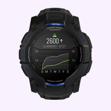 A Garmin Watches Instinct® 3 – 50 mm AMOLED 010-03020-03 in black displays a daily step count graph (2600 steps today), a weekly trend labeled S, M, T, W, T, F, S underneath, and shows a current total of 5721 steps.
