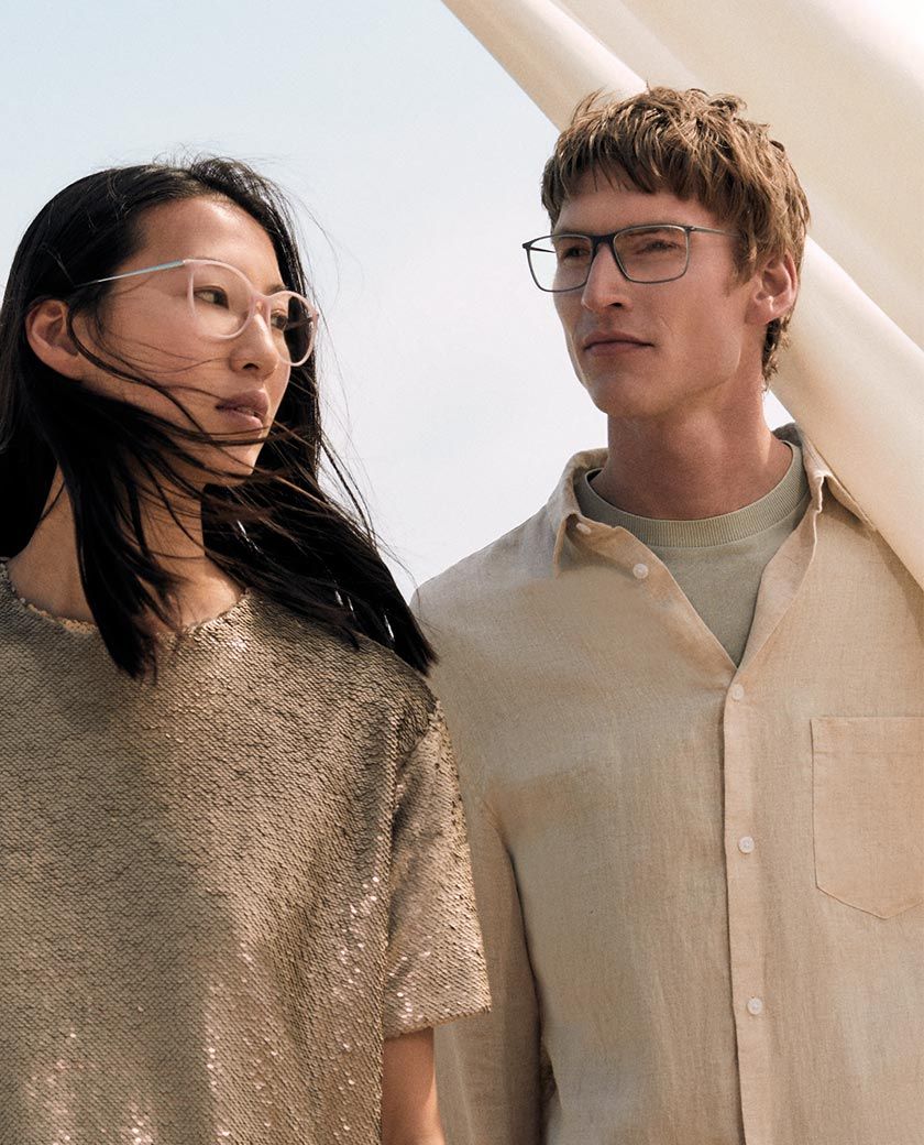 Two people wearing glasses stand side by side outdoors. The woman has long dark hair and wears a textured top, while the man wears a light button-up shirt. They look in different directions with a soft background behind them.