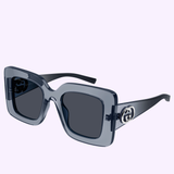 The Gucci Sunglasses GG2025S-003 49 feature square translucent gray frames, dark lenses, black arms, and a metallic logo on the temples—an iconic eyewear piece from Gucci Sunglasses, shown on a light lavender background.