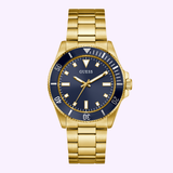 The Guess Watches GW0968G2 by Guess Watches is a men’s gold tone watch with a black dial, gold hour markers, gold metal link band, and a black bezel with white minute markings, all set against a light purple background.