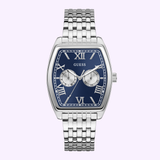 The Guess Watches GW0975G1 features a silver-tone recycled steel bracelet, rectangular blue face with Roman numerals and two subdials, shown against a light purple background.