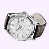 The Hamilton Jazzmaster Viewmatic Auto 40 mm Men Watch H32515555 by Hamilton Watches features a silver stainless steel case, white dial with a date display at 3 o’clock, and is complemented by a black leather strap. Shown on a light background, this automatic timepiece balances classic style with modern sophistication.