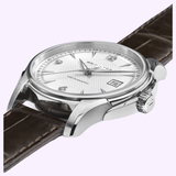 A close-up of the Hamilton Jazzmaster Viewmatic Auto 40 mm Men Watch H32515555 by Hamilton Watches, featuring a textured white dial with a date display, silver markers, and a brown leather strap. The elegant timepiece showcases a refined stainless steel case that enhances its sophisticated design, set against a light purple background.