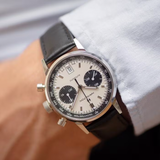 A close-up of a wrist wearing the Hamilton Watches American Classic Intra-Matic Auto Chrono 40 mm (H38416711), showing its white dial with black subdials, black leather strap, and silver case. The wearer has on a light blue shirt.