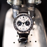 The Hamilton American Classic Intra-Matic Auto Chrono 40 mm Watch H38416711 by Hamilton Watches, featuring a white dial with two black subdials and tachymeter bezel on a black leather strap, is displayed on a metallic industrial surface.